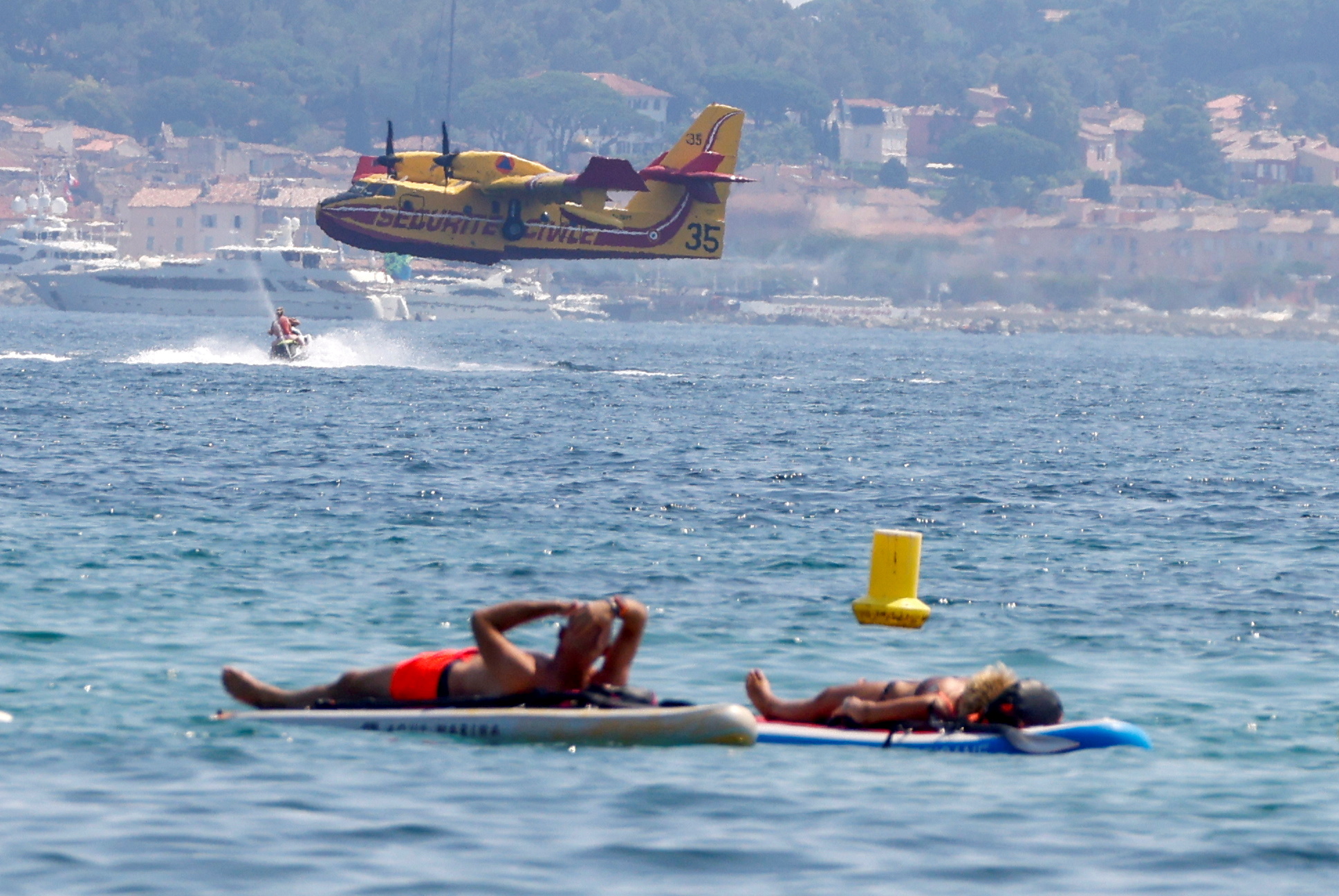 Las personas flotan en los tableros mientras un avión de Canadair vuela después de ser llenado con agua para ayudar con los esfuerzos para extinguir un gran incendio que estalló en la región de Var, en el Golfo de Saint Tropez, Francia el 17 de agosto de 2021. REUTERS / Eric Gaillard