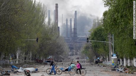 La gente camina con sus bicicletas por la calle mientras el humo se eleva sobre una planta de acero de Azovstal en Mariupol, Ucrania, el 2 de mayo.