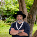 Un hombre de aspecto severo con una camiseta negra y un sombrero está de pie con los brazos cruzados frente a un árbol.