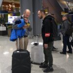 Dos jóvenes adultos miran un teléfono en un aeropuerto, con una maleta con ruedas a los lados.