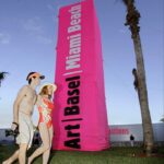 Un hombre y una mujer vestidos con trajes de baño pasan junto a una columna publicitaria rosa de Art Basel que sobresale del césped