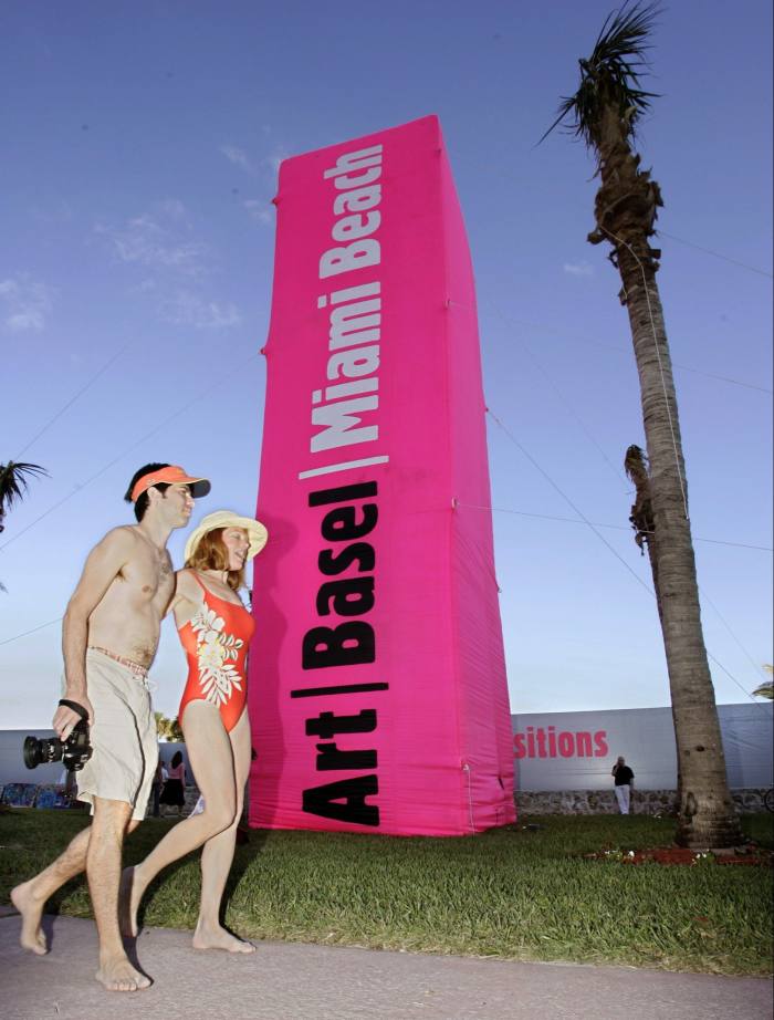 Un hombre y una mujer vestidos con trajes de baño pasan junto a una columna publicitaria rosa de Art Basel que sobresale del césped