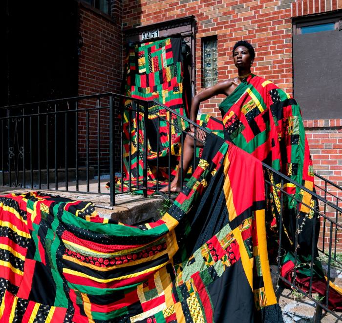 Foto de una mujer posada con orgullo envuelta en un tejido de retazos rojo, amarillo, verde y negro que desciende desde ella hasta el suelo y envuelve todo