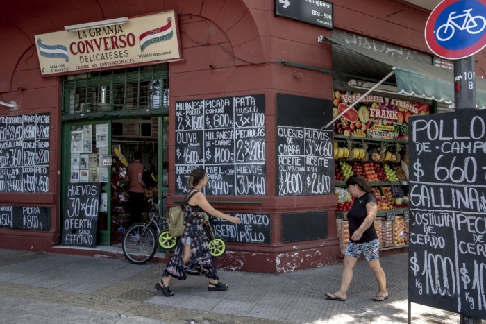 Las mujeres en una calle soleada pasan frente a una tienda de alimentos, donde hay varios precios escritos con tiza