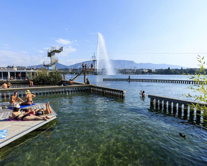 Bañistas en una plataforma de madera en el lago Bains des Pâquis de Ginebra, con plataformas de buceo al fondo