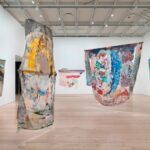 En una fotografía, cinco pinturas textiles abstractas y coloridas cuelgan del techo de una galería bien iluminada.