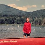 Una joven parada en una canoa roja en un lago con montañas detrás.