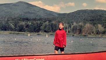 Una joven parada en una canoa roja en un lago con montañas detrás.