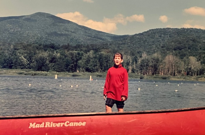 Una joven parada en una canoa roja en un lago con montañas detrás.