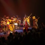 Una mujer con una túnica roja levanta sus manos alegremente rodeada en el escenario por músicos y bailarines con túnicas doradas.