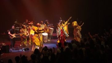 Una mujer con una túnica roja levanta sus manos alegremente rodeada en el escenario por músicos y bailarines con túnicas doradas.