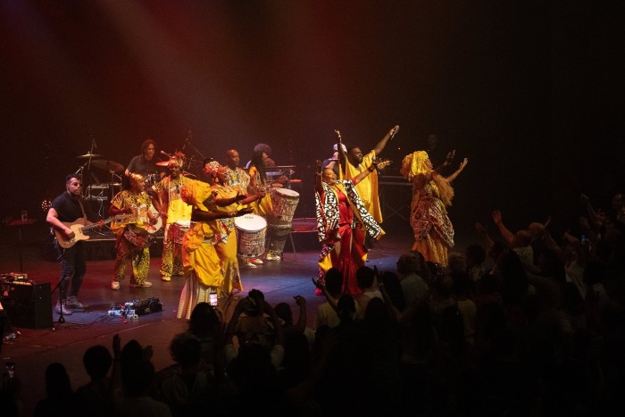 Una mujer con una túnica roja levanta sus manos alegremente rodeada en el escenario por músicos y bailarines con túnicas doradas.