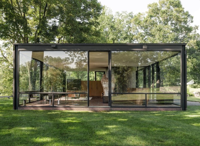 Una casa de cristal modernista situada en un paisaje verde y exuberante. La estructura tiene grandes ventanales de piso a techo en todos los lados.