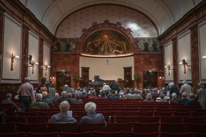 Personas llenando los asientos de terciopelo rojo de una pequeña sala de conciertos con decoraciones Art Nouveau.