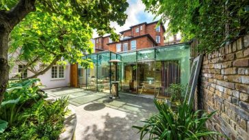 Una elegante ampliación en la planta baja y el patio trasero de una casa adosada del siglo XIX en Londres. La ampliación está hecha de vidrio con marcos de metal, mientras que el resto del edificio está hecho de ladrillo.