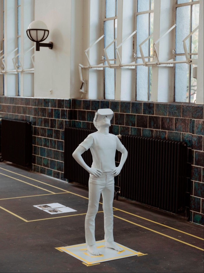 Escultura de yeso blanco en el suelo de un estudio de arte, que representa a un niño con las manos en las caderas, mirando hacia arriba y con gafas de realidad virtual.