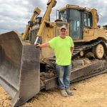 Bert Sterbutzel se encuentra junto a una gran topadora Caterpillar, vestido con una camisa amarilla neón, jeans y una gorra, en un sitio de construcción de tierra.