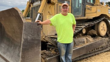 Bert Sterbutzel se encuentra junto a una gran topadora Caterpillar, vestido con una camisa amarilla neón, jeans y una gorra, en un sitio de construcción de tierra.