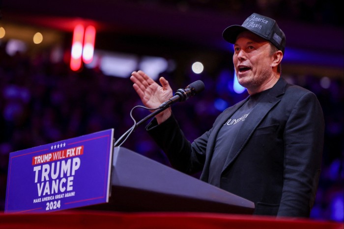 Elon Musk habla en un podio, vestido con una chaqueta y una gorra negras. El podio tiene un cartel que dice 'Trump Vance 2024'