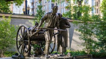 Una estatua de bronce de un hombre tirando de un carro cargado de pertenencias. Un niño se sienta en lo alto y una mujer camina a su lado.