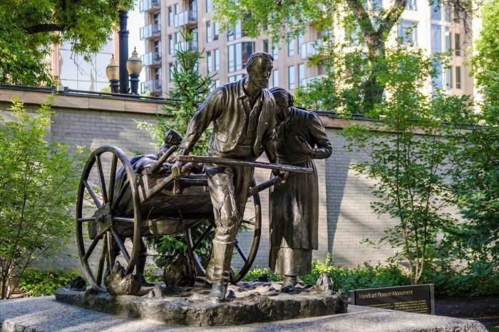 Una estatua de bronce de un hombre tirando de un carro cargado de pertenencias. Un niño se sienta en lo alto y una mujer camina a su lado.