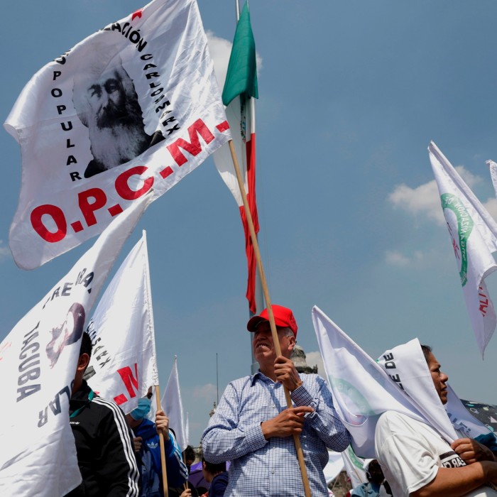 Los manifestantes en la Ciudad de México sostienen banderas y pancartas mientras se manifiestan contra la reforma judicial. Un hombre con gorra roja y camisa a cuadros sostiene de manera prominente una bandera, mientras otros se reúnen a su alrededor.