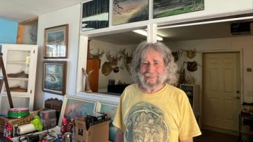 Un hombre se encuentra en una habitación que presenta pinturas de paisajes y cabezas de animales montadas.