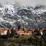 Un pintoresco albergue con arquitectura estilo chalet, ubicado entre árboles en la ladera de una montaña, enmarcado por espectaculares acantilados escarpados y picos nevados.