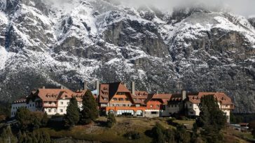 Un pintoresco albergue con arquitectura estilo chalet, ubicado entre árboles en la ladera de una montaña, enmarcado por espectaculares acantilados escarpados y picos nevados.