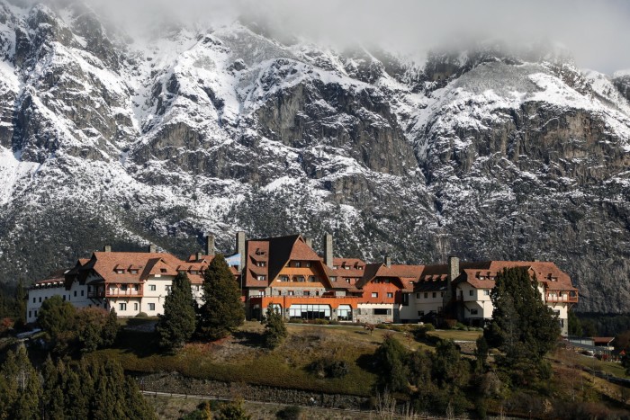 Un pintoresco albergue con arquitectura estilo chalet, ubicado entre árboles en la ladera de una montaña, enmarcado por espectaculares acantilados escarpados y picos nevados.