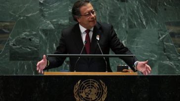 Un hombre vestido con traje oscuro, camisa blanca y corbata roja habla en un podio con el emblema de las Naciones Unidas frente a él, indicando que se dirige a una audiencia.