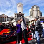 Una pareja se toma una selfie frente a un auto Red Bull de Fórmula 1 exhibido cerca del Caesars Palace, rodeado por una animada multitud.