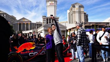 Una pareja se toma una selfie frente a un auto Red Bull de Fórmula 1 exhibido cerca del Caesars Palace, rodeado por una animada multitud.