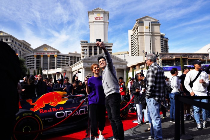 Una pareja se toma una selfie frente a un auto Red Bull de Fórmula 1 exhibido cerca del Caesars Palace, rodeado por una animada multitud.