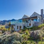 Una casa de lujo situada en una zona montañosa pintoresca bajo un cielo azul claro. La casa cuenta con grandes ventanales de vidrio y múltiples niveles.