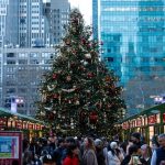 Un árbol de Navidad al final de un pasillo entre filas de puestos en Bryant Park Winter Village, con rascacielos detrás