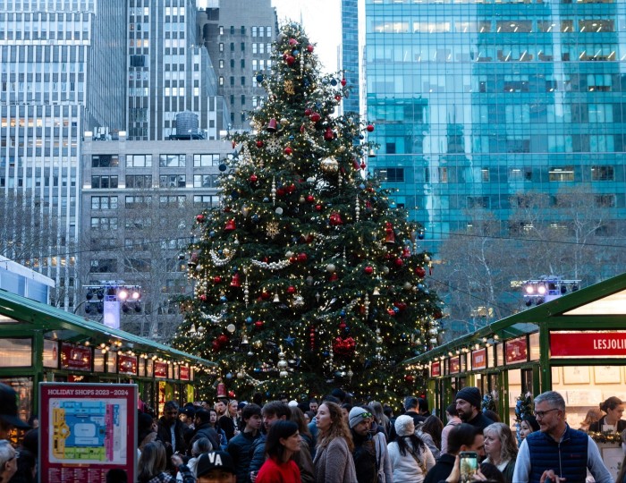 Un árbol de Navidad al final de un pasillo entre filas de puestos en Bryant Park Winter Village, con rascacielos detrás