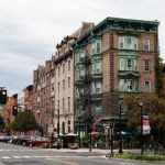 Una escena de una calle urbana donde la atención se centra en un edificio de esquina con adornos verdes ornamentados y una fachada de ladrillo. La calle tiene coches estacionados junto a la acera, algo de tráfico esperando en un semáforo en rojo, tiendas y peatones.