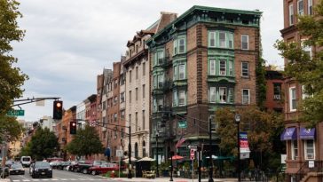 Una escena de una calle urbana donde la atención se centra en un edificio de esquina con adornos verdes ornamentados y una fachada de ladrillo. La calle tiene coches estacionados junto a la acera, algo de tráfico esperando en un semáforo en rojo, tiendas y peatones.