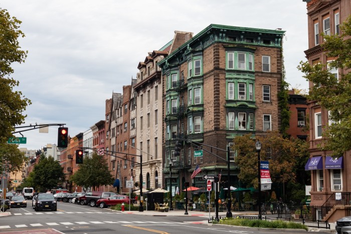 Una escena de una calle urbana donde la atención se centra en un edificio de esquina con adornos verdes ornamentados y una fachada de ladrillo. La calle tiene coches estacionados junto a la acera, algo de tráfico esperando en un semáforo en rojo, tiendas y peatones.