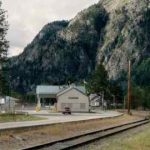 Un edificio blanco con cascada escrita en el frente. Hay un camión rojo frente al edificio. una línea de ferrocarril en primer plano y afloramiento de montaña rocosa en el fondo