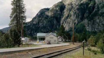 Un edificio blanco con cascada escrita en el frente. Hay un camión rojo frente al edificio. una línea de ferrocarril en primer plano y afloramiento de montaña rocosa en el fondo