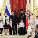 Nueve niños de distintas edades están de pie sobre una plataforma ornamentada con una mujer y dos hombres, con la bandera rusa al fondo.