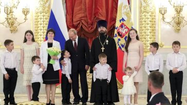 Nueve niños de distintas edades están de pie sobre una plataforma ornamentada con una mujer y dos hombres, con la bandera rusa al fondo.