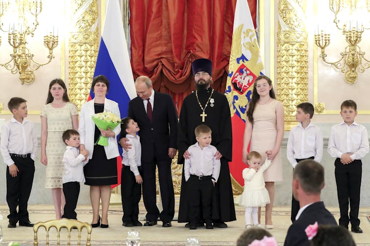 Nueve niños de distintas edades están de pie sobre una plataforma ornamentada con una mujer y dos hombres, con la bandera rusa al fondo.