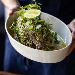 Un par de manos sosteniendo un medio pollo asado cubierto de hierbas en un plato de cerámica blanca en el restaurante Sailor