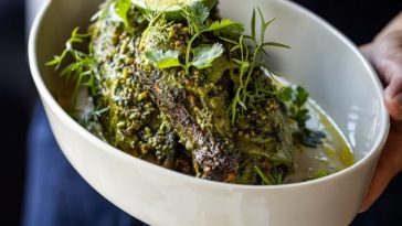 Un par de manos sosteniendo un medio pollo asado cubierto de hierbas en un plato de cerámica blanca en el restaurante Sailor