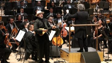 Un hombre se encuentra frente a una orquesta tocando una pequeña guitarra de cuatro cuerdas; junto a él, un hombre en un podio dirige