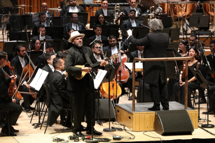 Un hombre se encuentra frente a una orquesta tocando una pequeña guitarra de cuatro cuerdas; junto a él, un hombre en un podio dirige