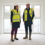 A man and a woman in hard hats and high-visibility vests survey a room in a newly built house